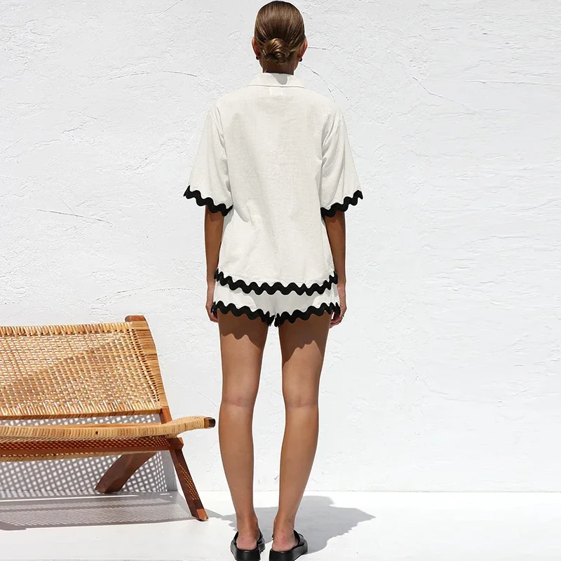 Woman wearing a white outfit with black trim standing in front of a white wall with a wicker chair.