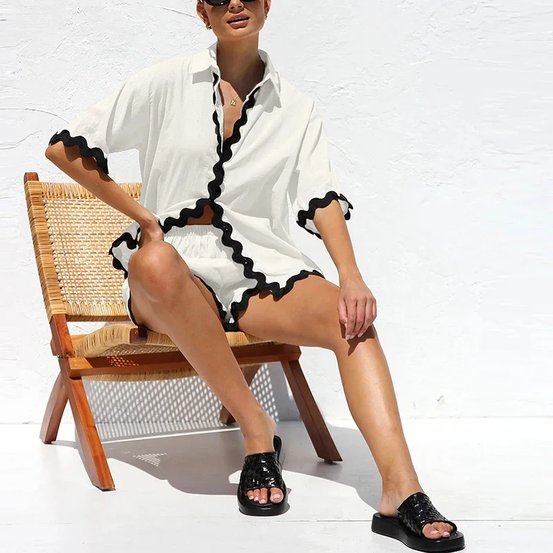 Woman in a white cover-up with black trim sitting on a wooden chair against a white wall.