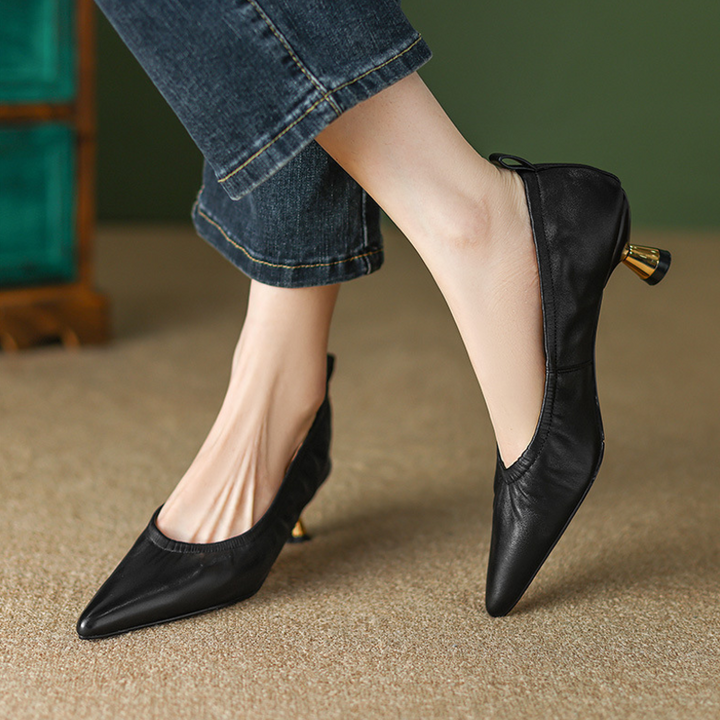 Black pointed-toe shoes worn with rolled-up jeans on a textured surface.