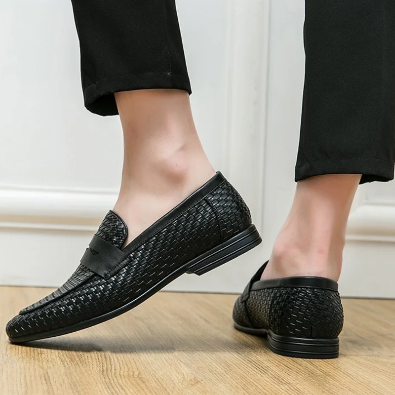 Black woven loafers worn indoors on a wooden floor.