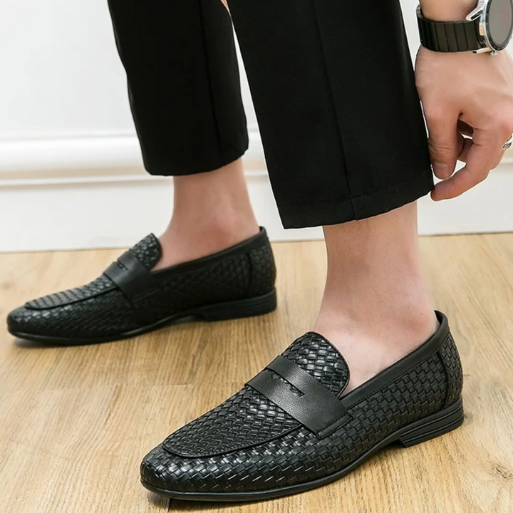 Black woven loafers worn with black pants on a wooden floor.