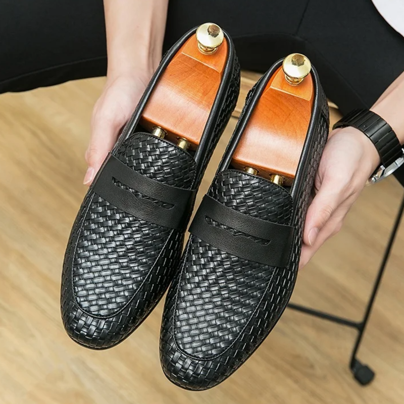 Black woven loafers with wooden soles held by a person on a wooden surface.