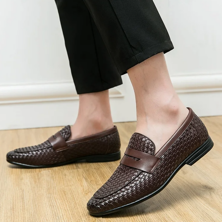 Brown woven loafers worn with black pants on a wooden floor.