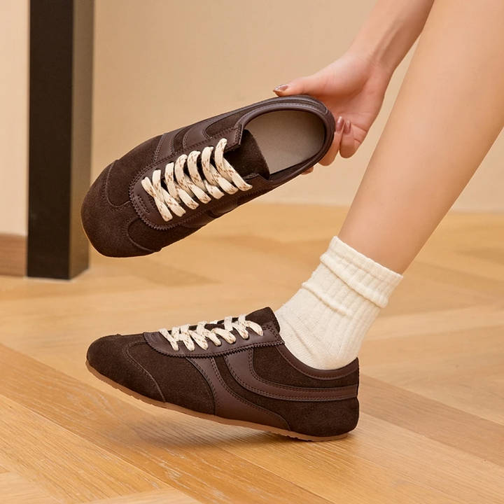 Brown slip-on shoes with white laces being held above a wooden floor.