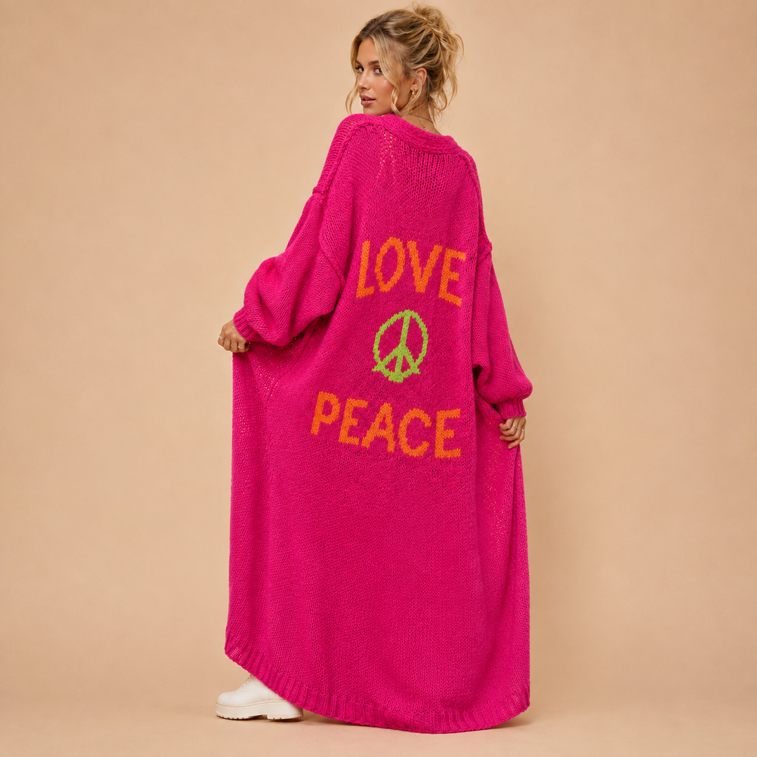 A woman wearing a bright pink chunky knit cardigan with 'LOVE PEACE' and a peace sign knitted into the back, shown from behind against a solid beige studio background.