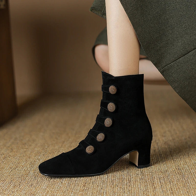 Black ankle boot with decorative buttons worn by a person on a textured surface.
