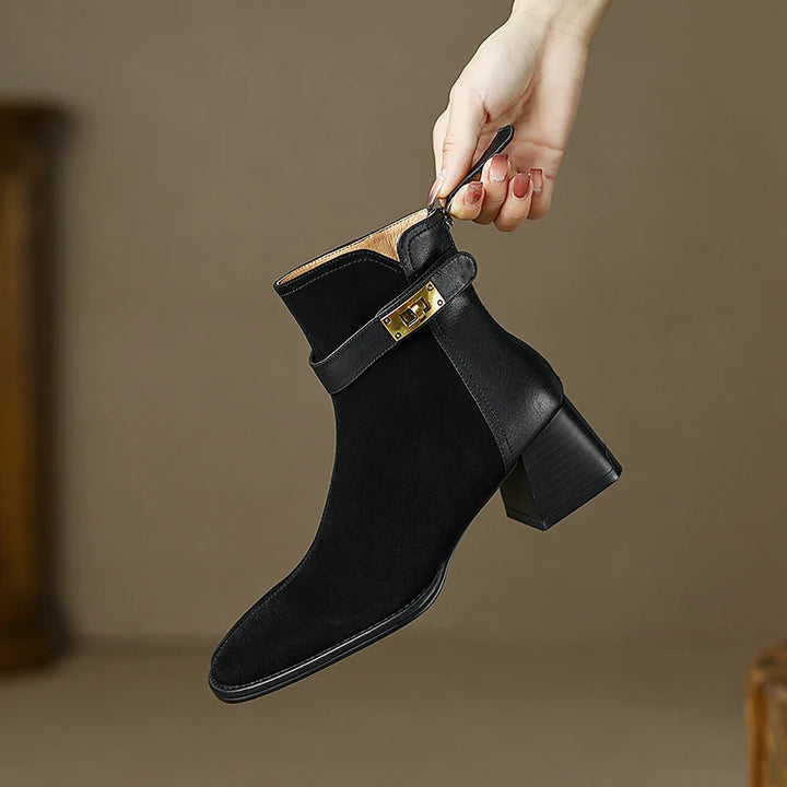 Black ankle boot with gold buckle held by a hand against a neutral background