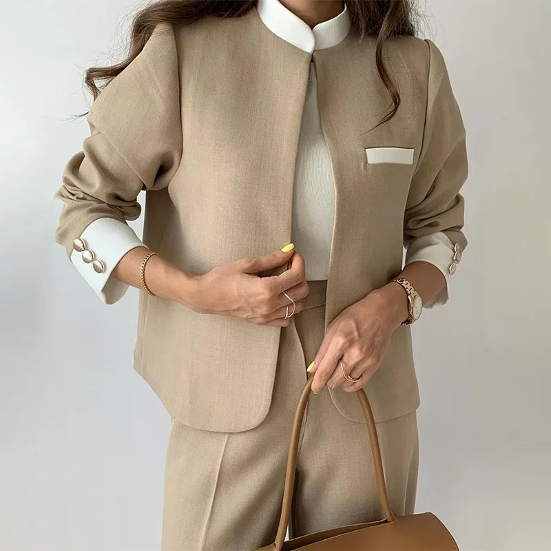 Person wearing a beige suit with a white shirt and holding a brown handbag on a plain background