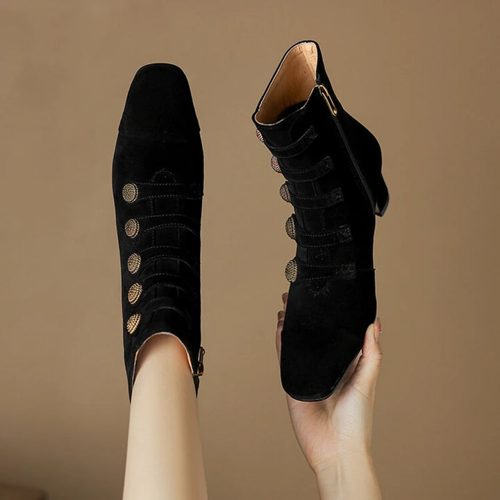 Black suede ankle boots with gold buttons held against a brown background