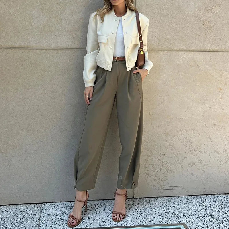 Woman wearing a white jacket, white shirt, and olive green pants against a beige wall.