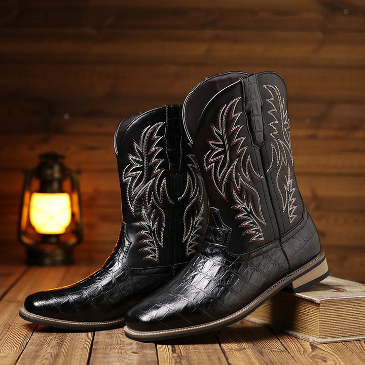 Black cowboy boots with white embroidery on a wooden surface with a lantern in the background