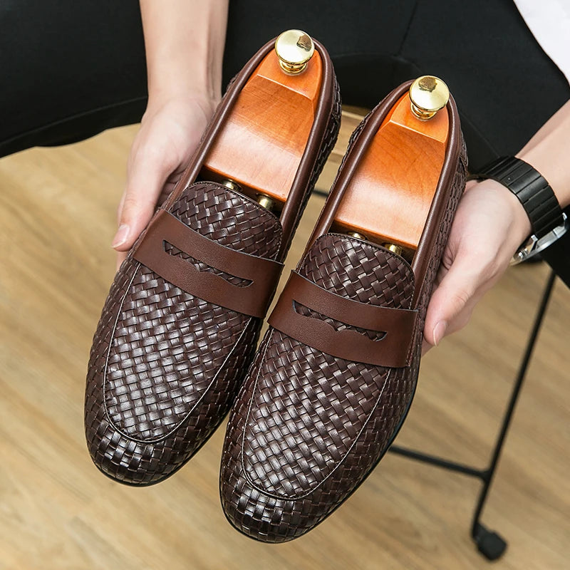 Brown woven loafers held by a person on a wooden floor.