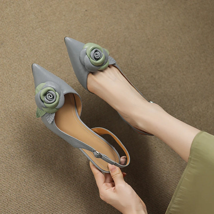 Gray high-heeled shoes with floral decorations held by a hand on a brown background