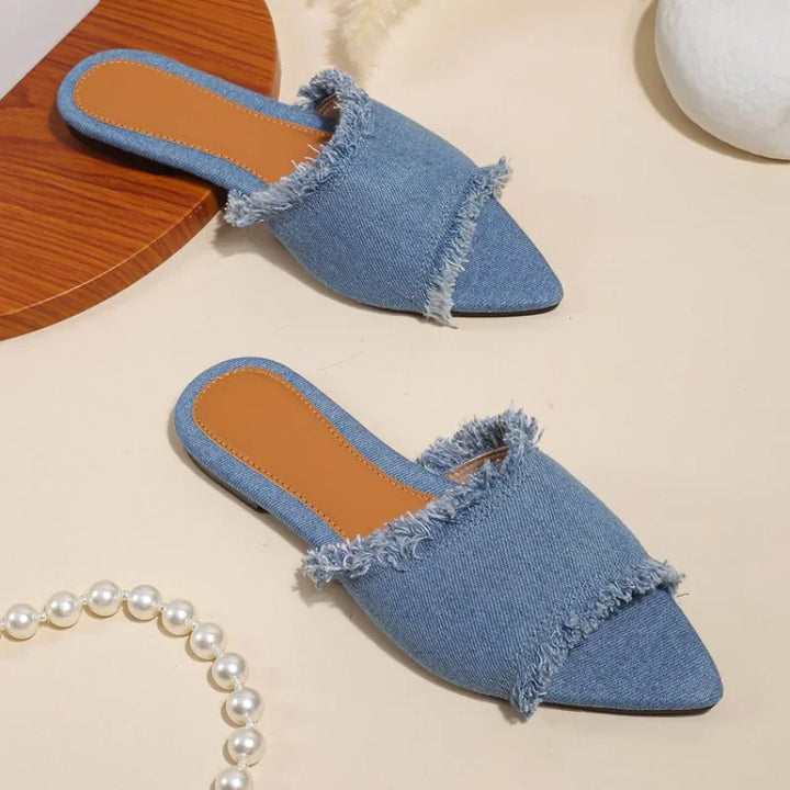 Pair of blue denim slippers on a beige surface with a pearl necklace.