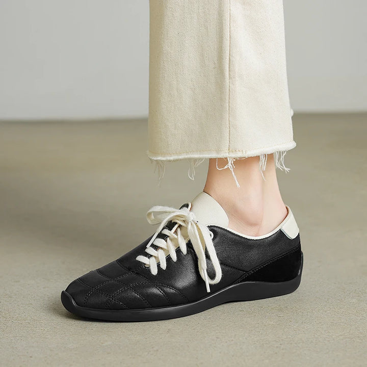 Black leather shoe with white laces worn with light-colored pants on a neutral background