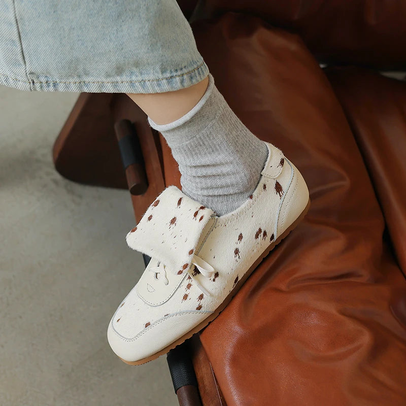 White sneakers with brown polka dots worn with gray socks on a brown leather chair.