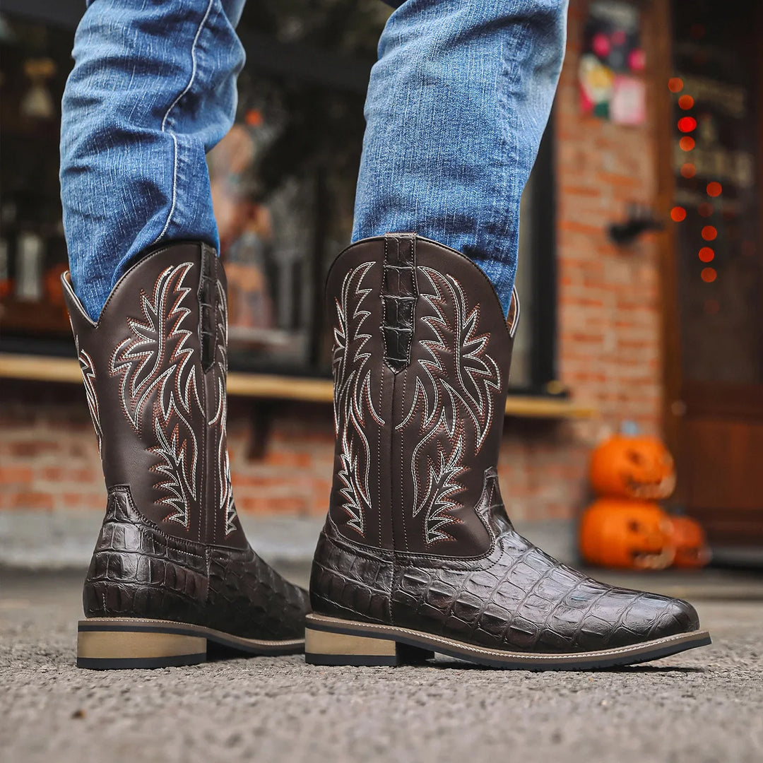 Brown leather cowboy boots with intricate designs worn by a person in blue jeans, with a blurred background.