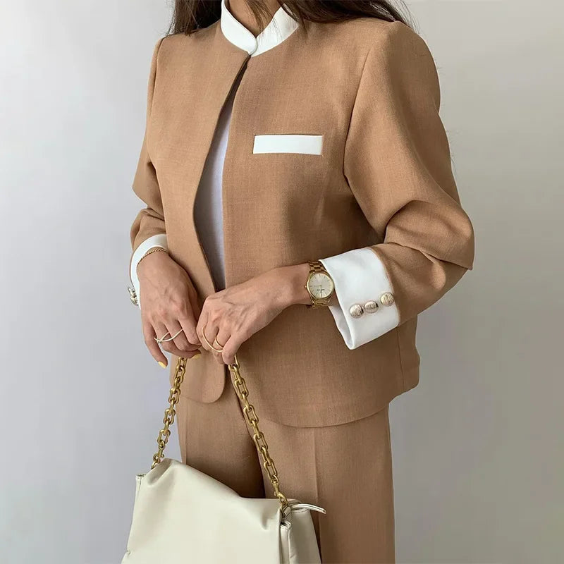 Person wearing a brown suit with white shirt and cuffs, holding a cream handbag against a neutral background.