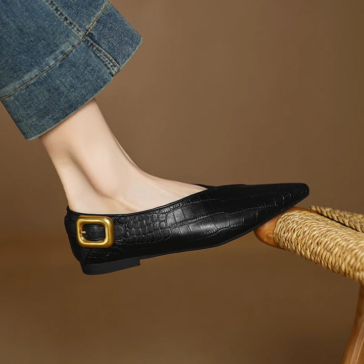 Black flat shoe with gold buckle worn by a person sitting on a wicker chair.