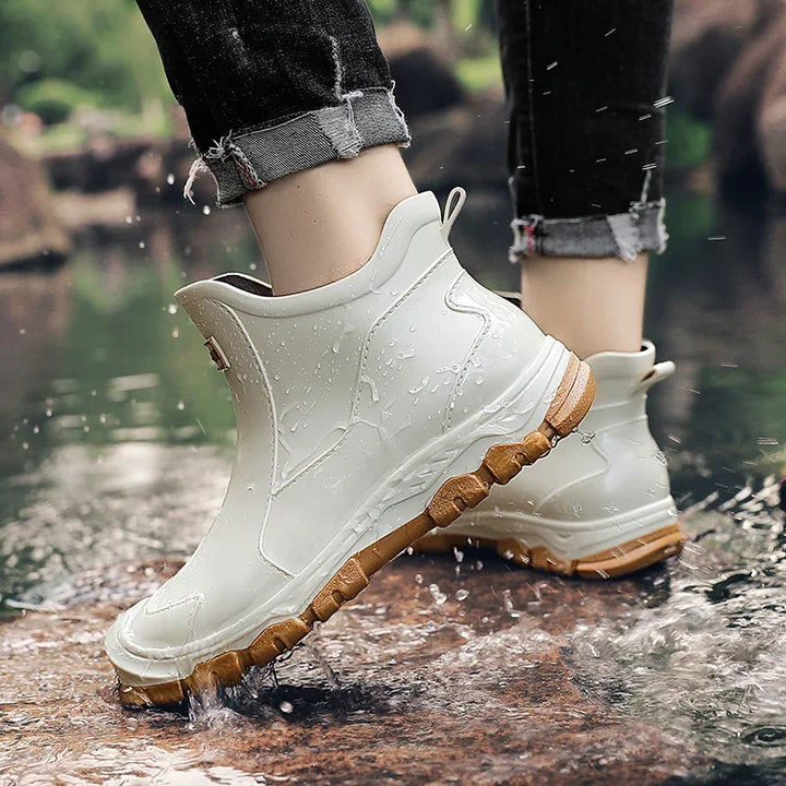 White rain boots with brown soles worn by a person stepping into water.