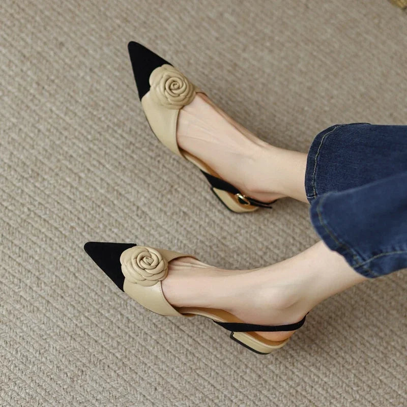 Beige high heels with black pointed toes and floral details worn by a person on a textured floor.
