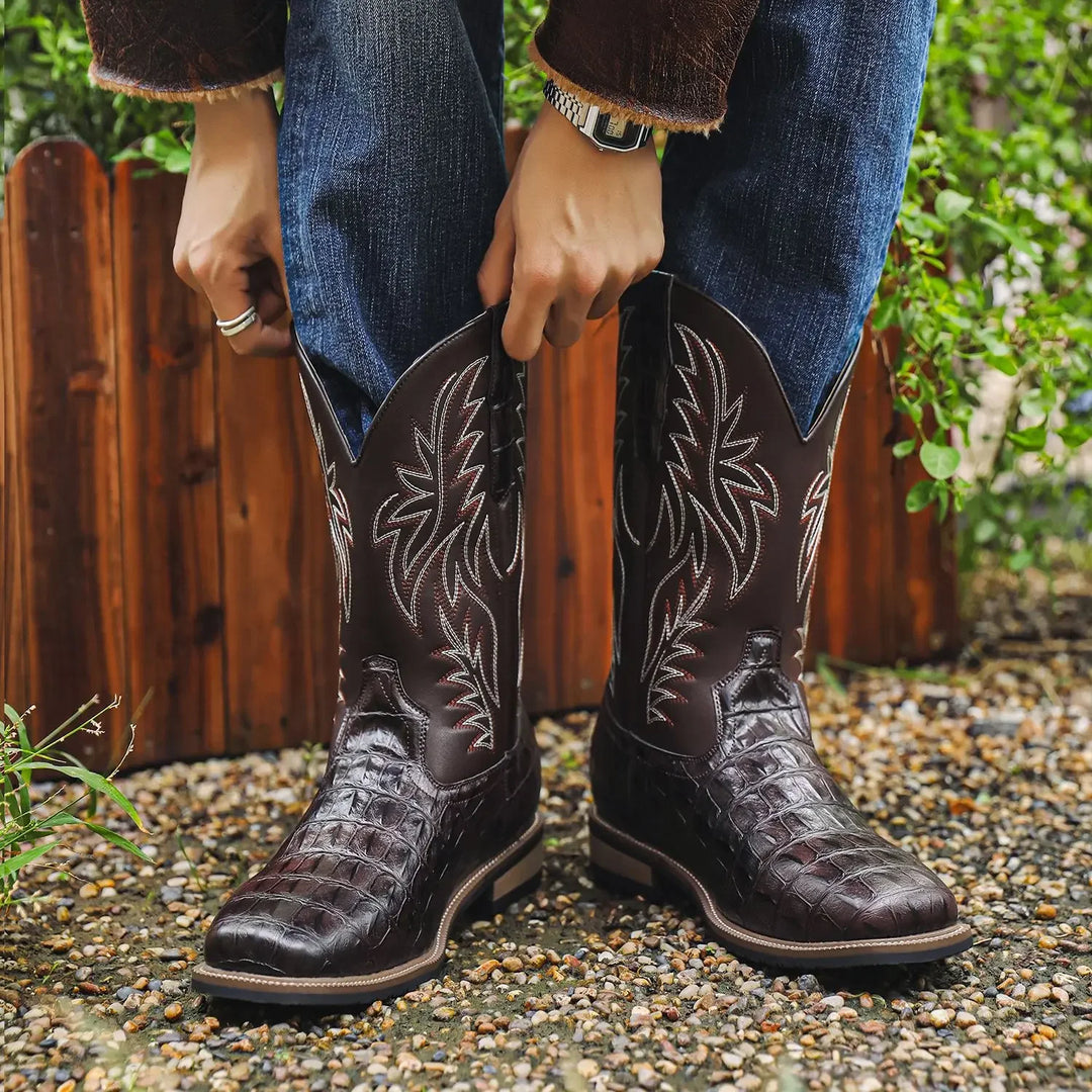 Person wearing dark brown cowboy boots with intricate designs, standing on a gravel surface with a wooden fence and greenery in the background.