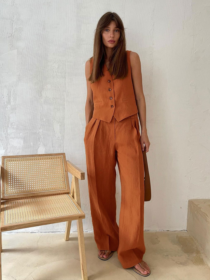 Woman wearing a brown sleeveless top and wide-leg pants standing next to a wicker chair against a white wall.