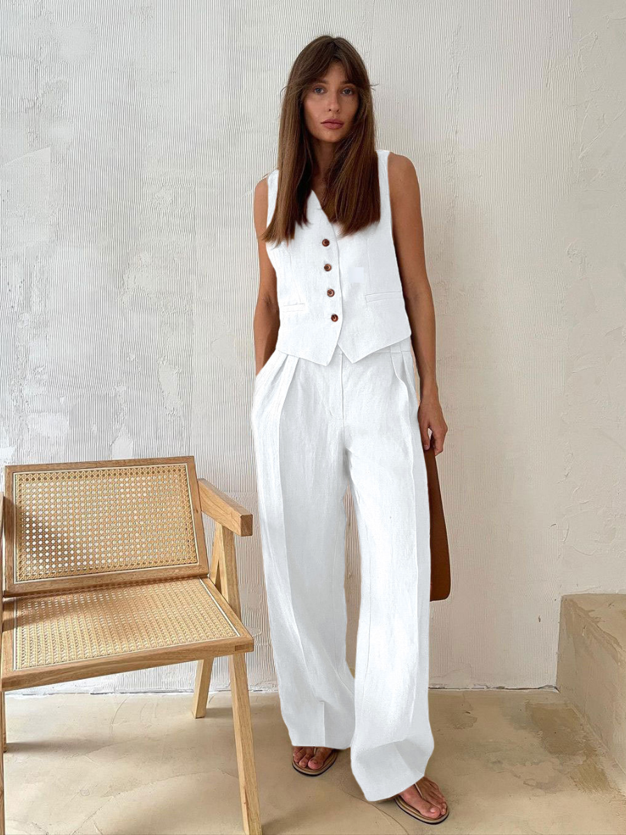 Woman in a white sleeveless top and wide-leg pants standing next to a wooden chair against a light-colored wall.