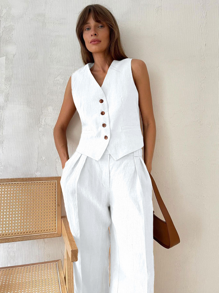 Woman wearing a white vest with buttons and white pants, standing against a neutral wall.