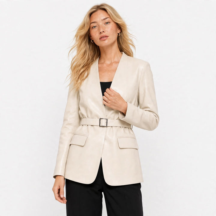 A woman modeling a light beige leather blazer with a matching belt and silver buckle, worn over a black top and black trousers, against a plain white studio background.