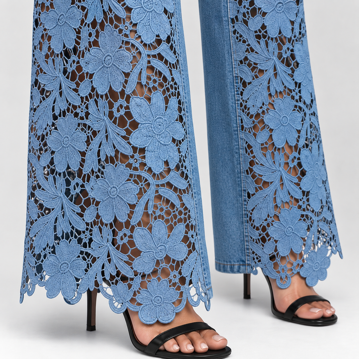 Close-up of a person wearing wide-leg blue jeans with a floral lace overlay and black heeled sandals against a light gray studio background.