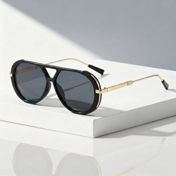Black aviator sunglasses with gold metal frames and dark gray lenses, resting on a white rectangular block against a light gray background.