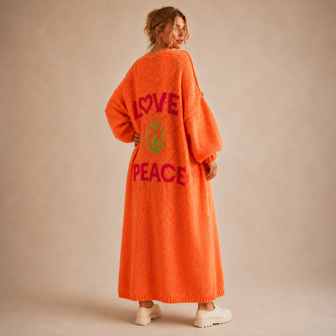 A woman stands with her back to the camera wearing a bright orange fuzzy cardigan with 'LOVE PEACE' and a peace sign graphic in pink and green on the back, paired with white sneakers against a beige studio backdrop.