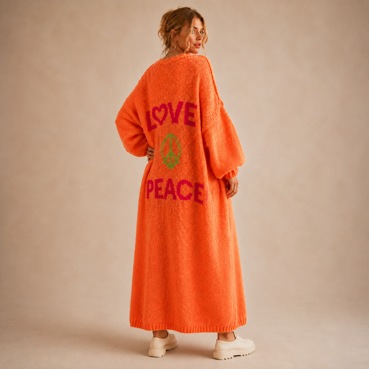 A woman stands with her back to the camera wearing a bright orange fuzzy cardigan with 'LOVE PEACE' and a peace sign graphic in pink and green on the back, paired with white sneakers against a beige studio backdrop.
